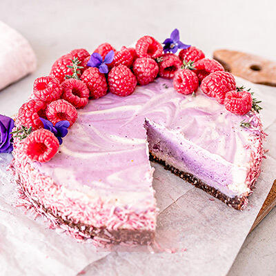 Raw Vegan Cheesecake Roh-veganer Cheesecake mit cremiger, violett-weiß marmorierter Füllung und dunklem Boden. Eine Scheibe ist herausgeschnitten. Oben liegt eine Dekoration aus frischen Himbeeren und lila Blüten.