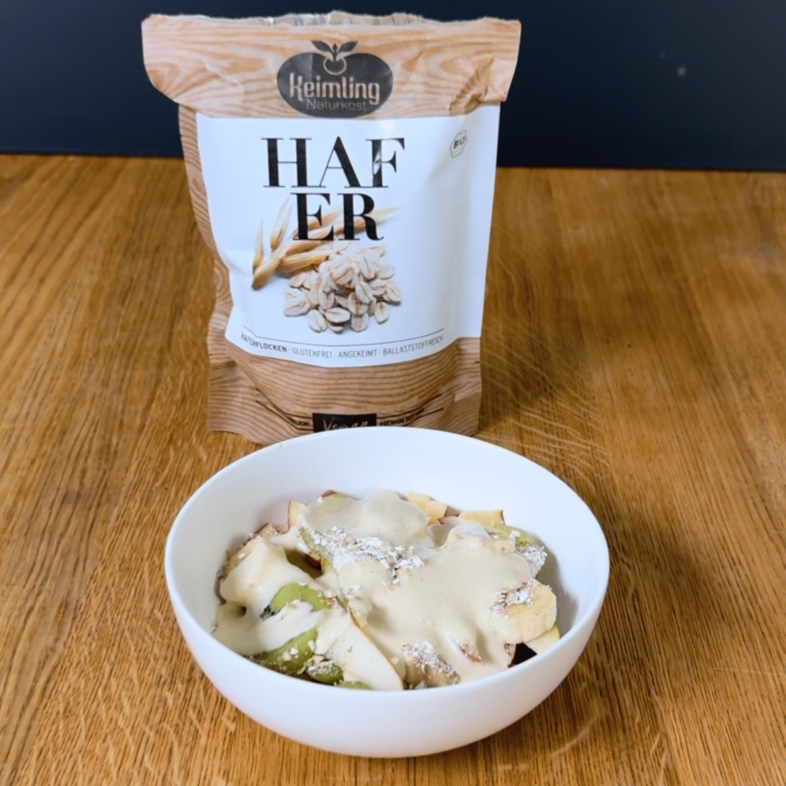 Müsli Superfood Mandeldrink Eine weiße Schüssel mit Müsli, belegt mit Bananen, Kiwi und einer weißen Mandelmus-Creme. Im Hintergrund steht eine Packung Keimling Naturkost Haferflocken.