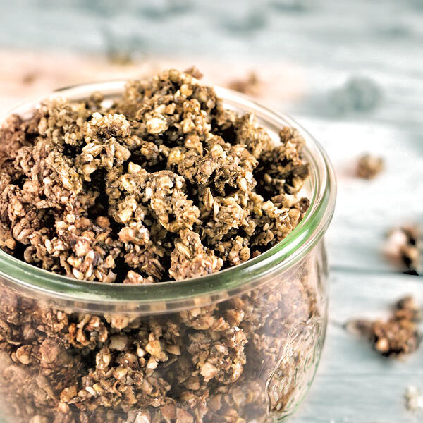 Spekulatius-Schoko Granola, roh-vegan Ein Glas ist gefüllt mit braunem Spekulatius-Schoko Granola aus Mandel- und Haselnussstücken. Granola liegt auch verstreut auf einer hellen Holzoberfläche.