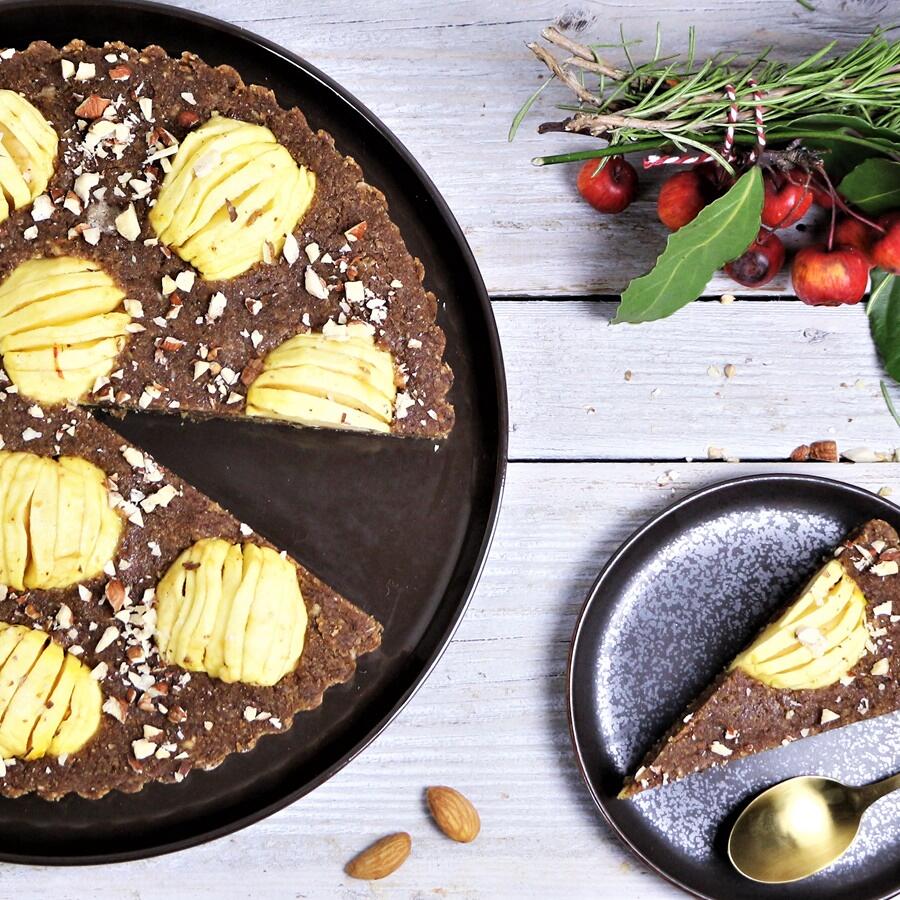 Bratapfelkuchen mit Haselnüssen, roh-vegan Roh-veganer Bratapfelkuchen mit Haselnüssen auf dunklem Teller, ein Stück Kuchen auf kleinem Teller neben goldenem Löffel, Dekoration mit kleinen roten Äpfeln und grünen Zweigen.