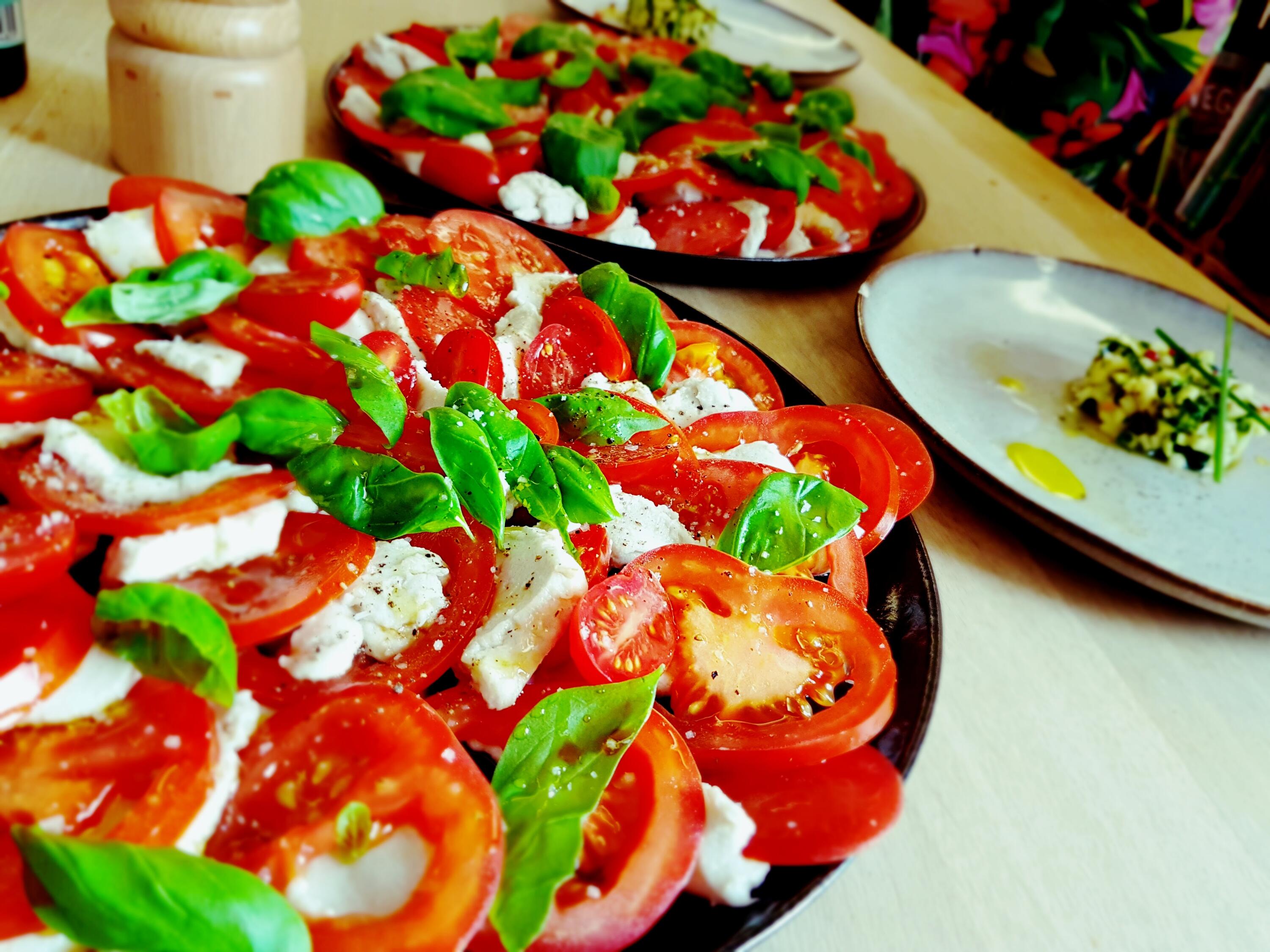 Cashew Kokos Mozzarella Zwei Teller mit veganem Cashew Kokos Mozzarella, garniert mit Tomatenscheiben, frischen Basilikumblättern und Gewürzen stehen auf einem Holztisch. Ein weiterer Teller mit einem kleinen Kräutergericht ist im Hintergrund zu sehen.