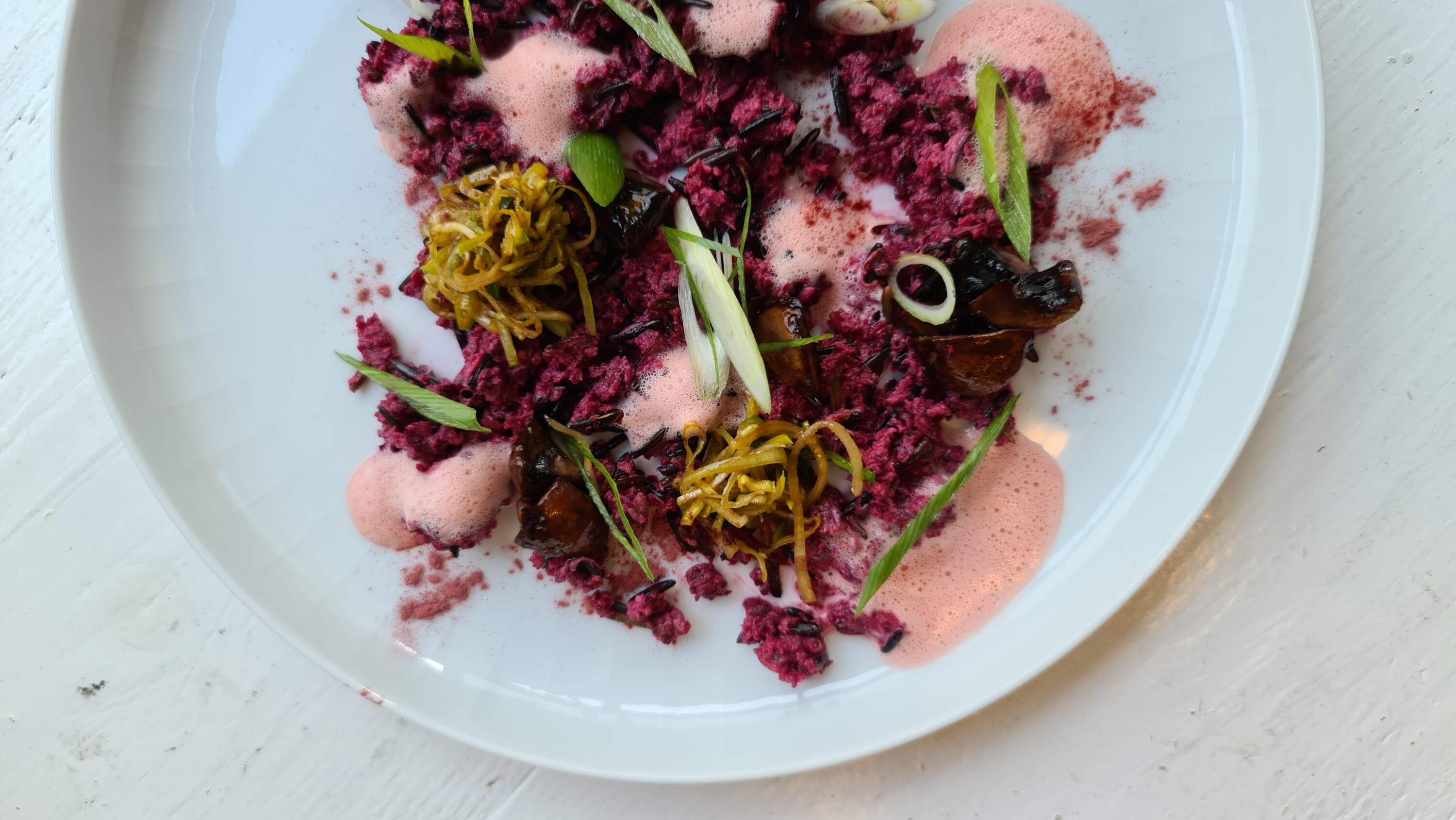 Rote Beete Festtags Risotto mit gekeimtem Wildreis Auf Weißem Teller liegt ein Gericht mit roter, körniger Masse, rosa Schaum, braunen Pilzen und grünen Frühlingszwiebelringen, garniert mit dünnen, goldgelben Gemüsestreifen.