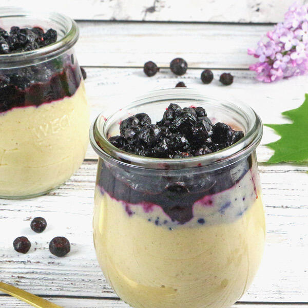 Köstlicher Sesam-Pudding, roh-vegan Zwei Gläser mit cremigem Sesam-Pudding, das obere Drittel ist mit dunklen Waldheidelbeeren bedeckt. Der Pudding steht auf einem weißen Holztisch, daneben liegen einzelne Beeren und eine grüne Blattranke.