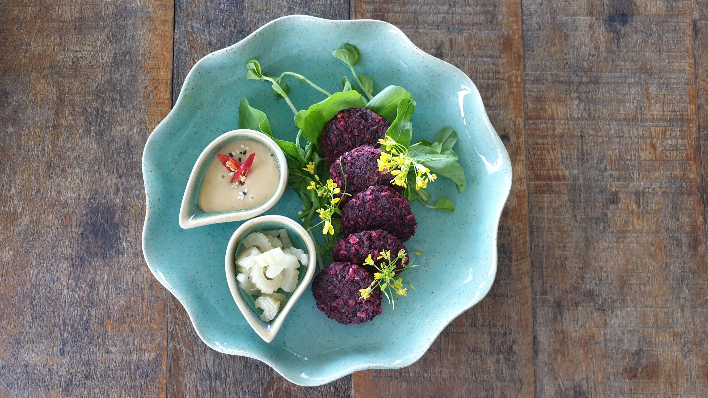 Rote Beete Zitronengras Burger mit Chili Mayo und Fermentiertem Sellerie Blaue Keramikplatte mit vier kleinen, runden, lila gebratenen Gemüse-Patties, grünen Blättern und gelben Blüten. Zwei kleine Schalen enthalten helle Soße mit Chilistücken und geschnittenen Selleriestücken.