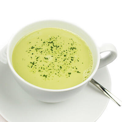 Eine weiße Tasse gefüllt mit cremigem, hellgrünem Matcha Cappuccino, bestreut mit grünen Matcha-Pulverflocken, steht auf einer weißen Untertasse mit einem Löffel.