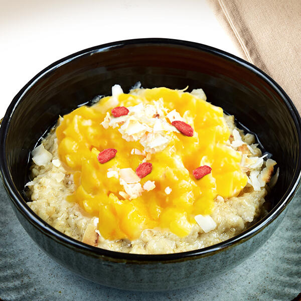 Rohkost Mango Porridge Schwarze Schüssel mit Mango Porridge aus Haferflocken, garniert mit leuchtend gelber Mangosoße, roten Gojibeeren und weißen Kokosraspeln, stehend auf grauem Untergrund neben einem hellbraunen Tuch.