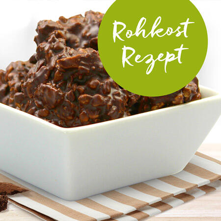 Weiße Schüssel mit mehreren unregelmäßigen Schokoladen-Häufchen. Grüner Kreis mit weißer Schrift Rohkost Rezept über den Häufchen. Schüssel steht auf gestreifter Unterlage.