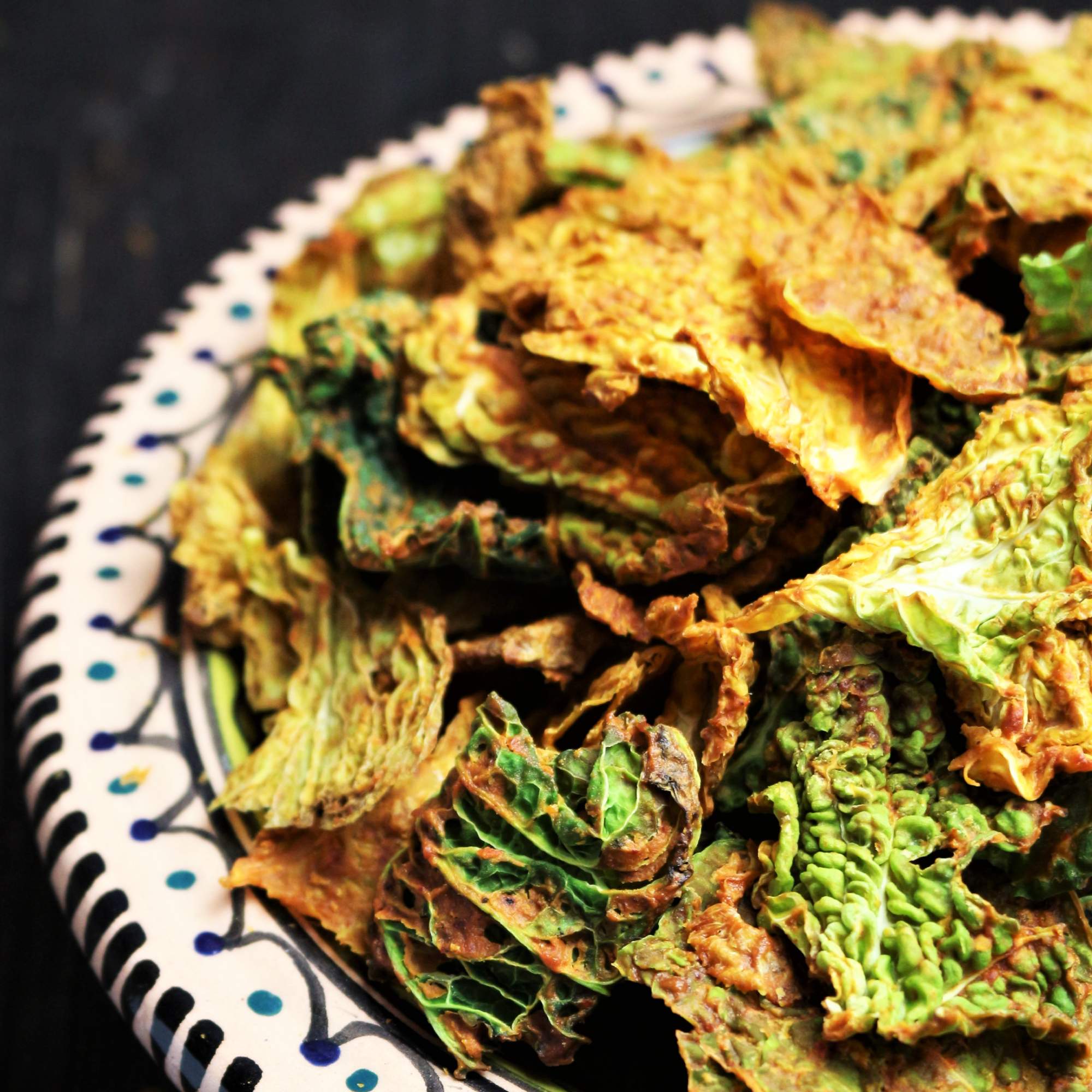 Chipotle Chips Knusprige Wirsingchips sind in einem weißen Teller mit blauen und schwarzen Mustern angerichtet. Die Chips zeigen verschiedene Grüntöne und sind leicht gebräunt.