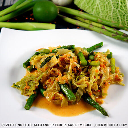 Wirsingcurry Gedünsteter Wirsing und grüner Spargel werden mit einer cremigen, orangen Sauce auf einem weißen Teller serviert. Im Hintergrund sind frische Spargelstangen, Limette und Wirsingblätter zu sehen.