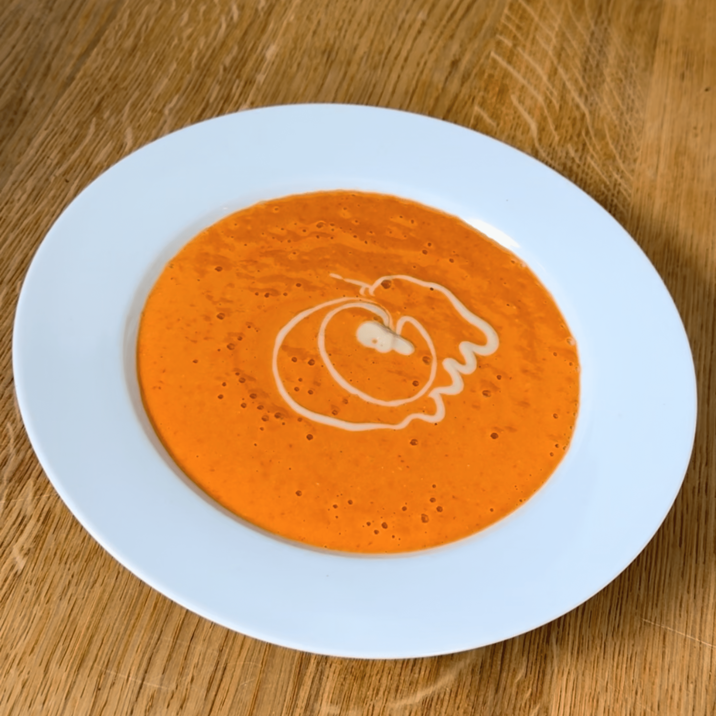 Karotten Creme Suppe Weiße Schale mit orangefarbener Karotten-Cremesuppe. Oben auf der Suppe ist ein geschwungener, weißer Sahne- oder Mandelpüree-Dekor in spiralförmigem Muster. Schale steht auf holzfarbenem Tisch.