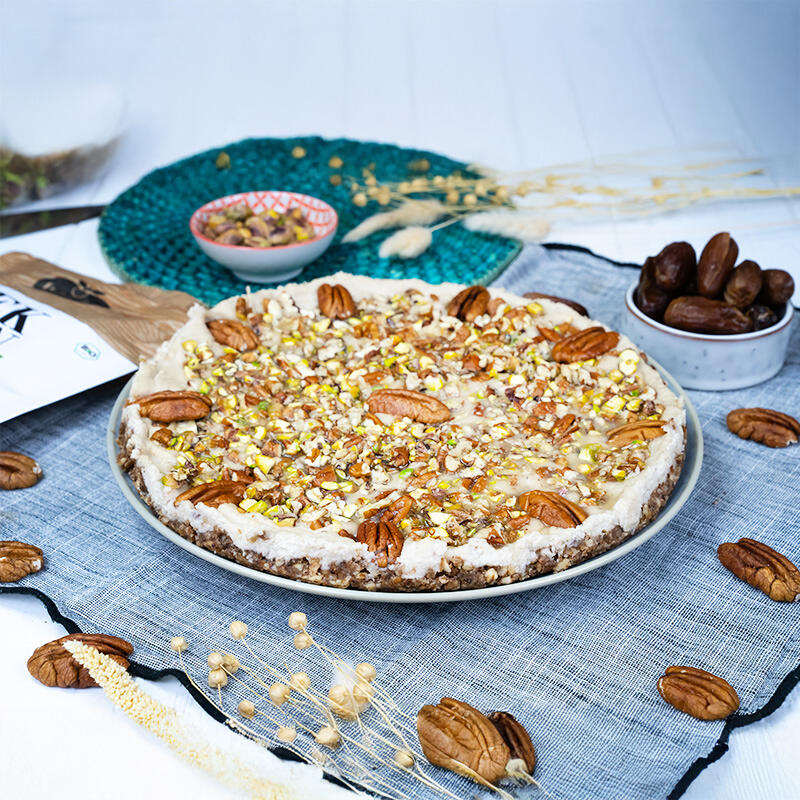 Pekanuss Schnitten Raw & vegan von Deutschland is(s)t vegan Runde Pekanuss-Schnitten mit nussigem Boden und cremiger Cashew-Schicht. Das Topping besteht aus Pekannüssen und gehackten Pistazien. Im Hintergrund liegen Datteln und weitere Pekannüsse.