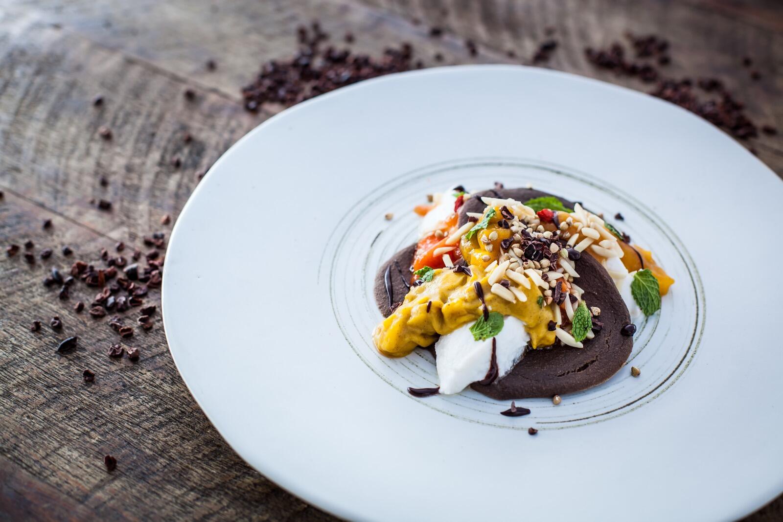 Bananen Pancakes Auf einem weißen Teller ist ein dekorativ angerichteter Teller mit einer braunen Creme, weißen Stücken, gelber Sauce, gehackten Nüssen und Minzblättern zu sehen.