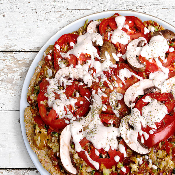 Raw-Pizza von Peter Dreverhoff Roh-vegane Pizza von Peter Dreverhoff liegt auf einem weißen Teller. Sie ist belegt mit Tomatenscheiben, Champignons, Nüssen, Gemüse und einem weißen, cremigen Dressing.