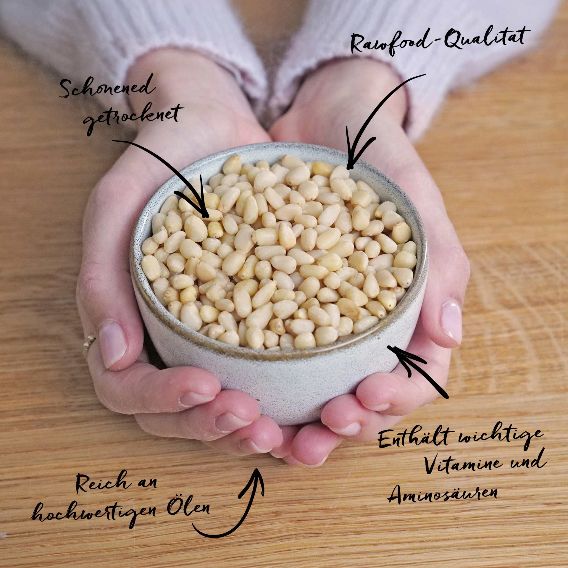 Zedernkerne, bio 100 g Hände halten eine kleine Schale mit geschälten Pinienkernen auf einem Holzuntergrund. Pfeile und Schrift beschreiben die Pinienkerne als Schonend getrocknet, Rawfood-Qualität, Reich an hochwertigen Ölen und Enthält wichtige Vitamine und Aminosäuren.