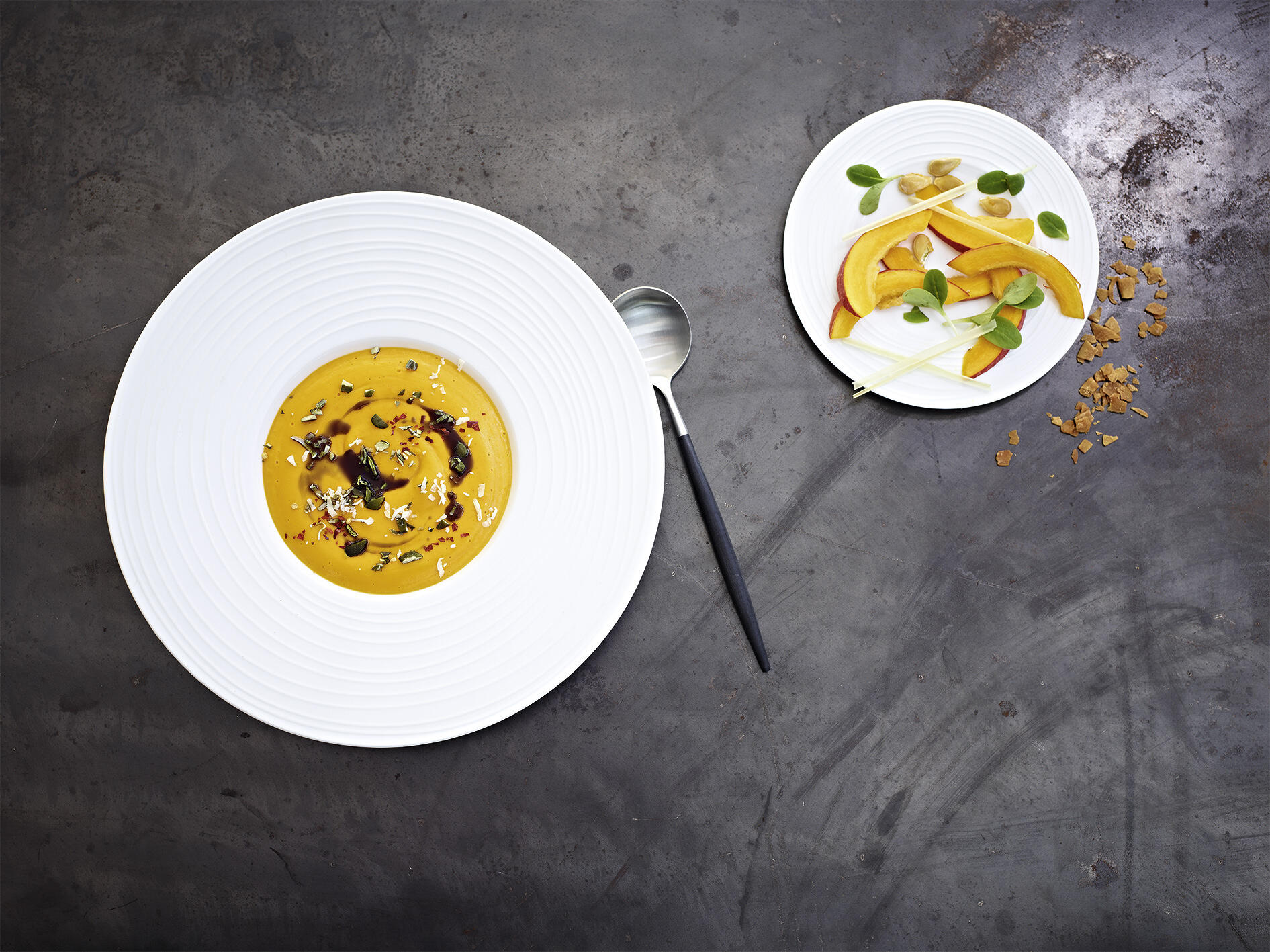 Kürbis-Kokos-Suppe mit Zitronengras Weiße tiefe Teller mit cremiger Kürbis-Kokos-Suppe, garniert mit Kürbiskernen, Gewürzen und Kräutern. Daneben ein kleiner Teller mit Zitronengras, Kürbisspalten und Blättern, plus Löffel.