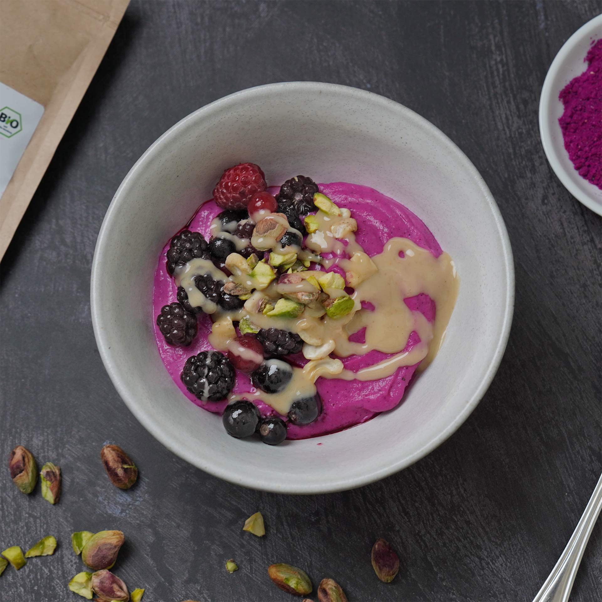 Winterzauber Dragonfruit Smoothie Bowl Eine weiße Schale mit cremiger, pinkfarbener Dragon Fruit Smoothie Bowl. Oben liegen Himbeeren, Brombeeren, Blaubeeren, gehackte Pistazien und eine beige cremige Sauce. Mehrere Pistazien liegen um die Schale herum auf einer dunklen Oberfläche.