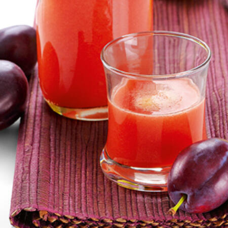 Ein Glas und eine Karaffe mit frisch gepresstem, rotem Pflaumensaft stehen auf einer violetten Unterlage. Zwei ganze Pflaumen liegen daneben.