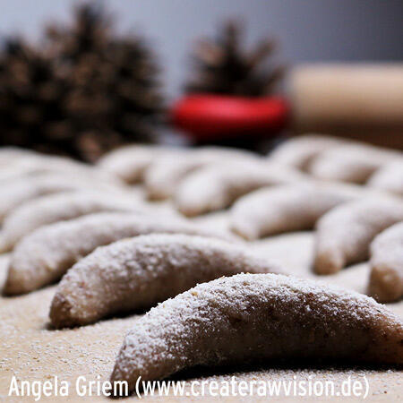 Mehrere halbmondförmige Rohkost-Pralinen liegen auf einer bemehlten Arbeitsfläche, bestäubt mit Mehl, im unscharfen Hintergrund sind Tannenzapfen und ein rotes Objekt zu erkennen.