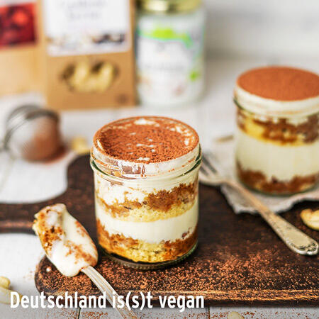 Gläser mit Schichtdessert aus veganem Biskuit, Creme und Kakaopulver stehen auf einem dunklen Untergrund. Ein Löffel mit Creme liegt neben einem Glas.