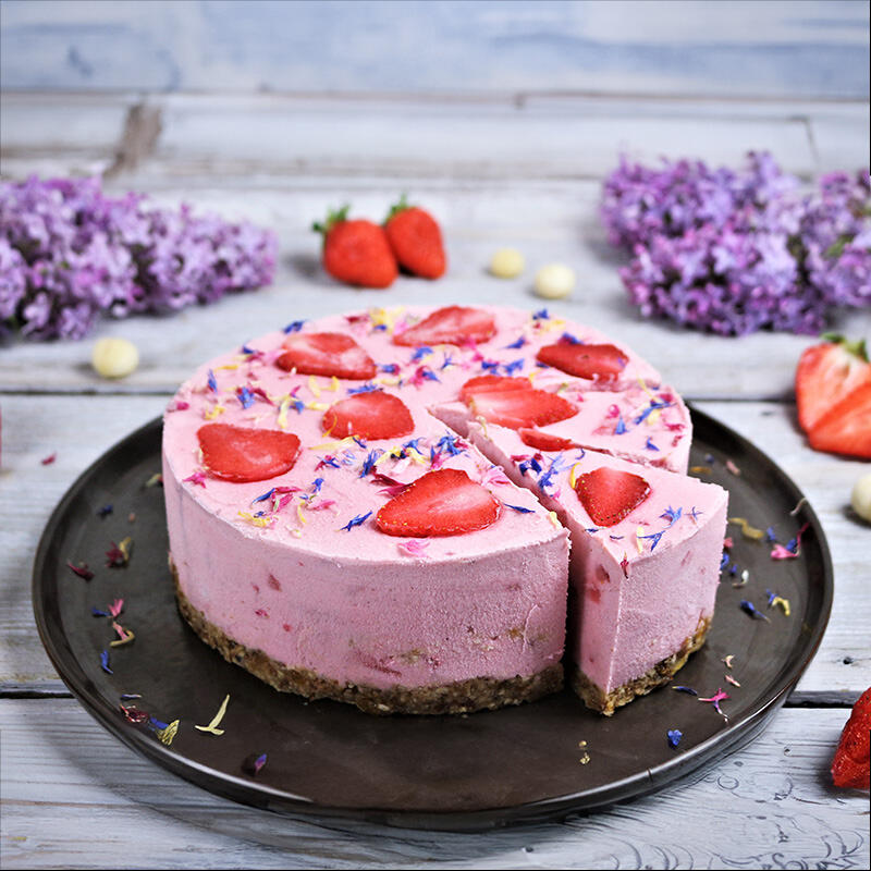Feinster Macadamia-Erdbeeren Cheesecake Runder, rosa Macadamia-Erdbeeren Cheesecake mit Mandelboden steht auf einem dunklen Teller. Ein Stück ist herausgeschnitten. Die Oberfläche ist mit Erdbeerscheiben und bunten Blüten verziert.