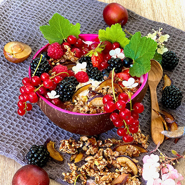 Knuspriges Pflaumen-Granola Schale mit Knusprigem Pflaumen-Granola, garniert mit roten Johannisbeeren, Brombeeren, Himbeeren, grünen Blättern und weißen Blüten, auf grauem Tuch mit Holzlöffeln und frischen Pflaumen.