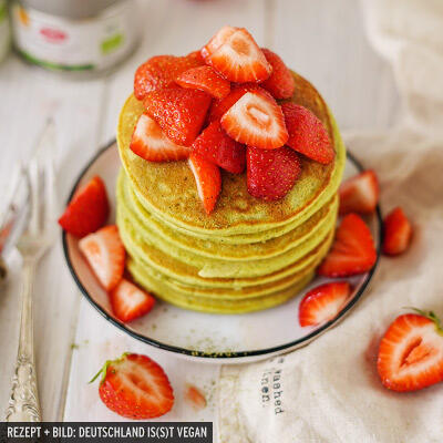 Ein Stapel grüner Pancakes auf einem weißen Teller mit schwarzem Rand. Oben und rundherum liegen frische, rote Erdbeeren, einige halbiert, als Topping.