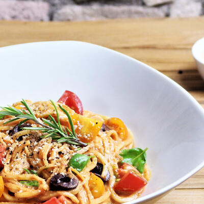 Spaghetti in Tomatencremesoße mit Oliven und Parmesan, roh-vegan Ein Teller mit Spaghetti aus rohem Kohlrabi in cremiger Tomatensoße, garniert mit schwarzen Oliven, halbierten roten und gelben Cocktailtomaten, frischen Basilikumblättern, rohem Parmesan und Rosmarinzweigen.