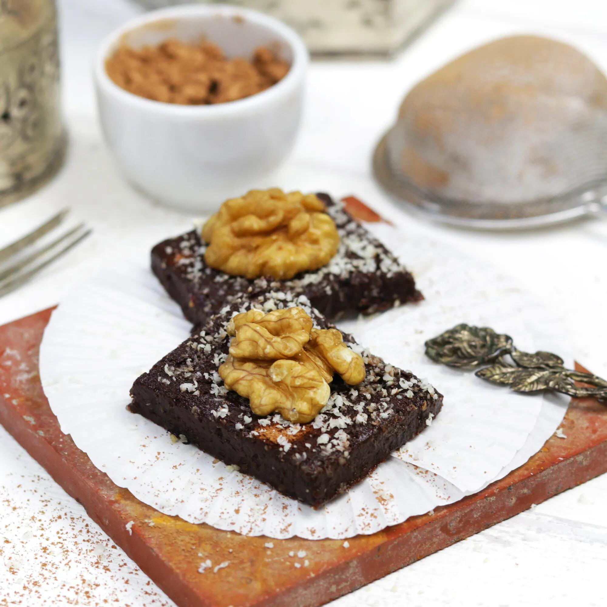 Rohkost-Brownies für Anfänger Zwei quadratische Rohkost-Brownies mit Walnusskernen und feinen Nussraspeln liegen auf weißen Papierförmchen auf einer roten Platte. Im Hintergrund stehen eine weiße Schale mit Nüssen und ein Sieb.