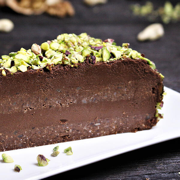 Death by Chocolate Pistazientorte, roh-vegan Ein großes Stück roh-vegane Schokoladentorte mit zwei feinen Schoko-Biskuit- und zwei Schoko-Cremeschichten. Die Torte ist mit gehackten Pistazien oben und außen am Rand bedeckt.