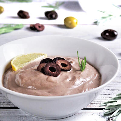 Weiße Schüssel mit cremigem Olivendip, garniert mit schwarzen Olivenringen, einem Zitronenschnitz und einem kleinen Rosmarinzweig auf hellem Untergrund. Im Hintergrund liegen verstreut schwarze und grüne Oliven.