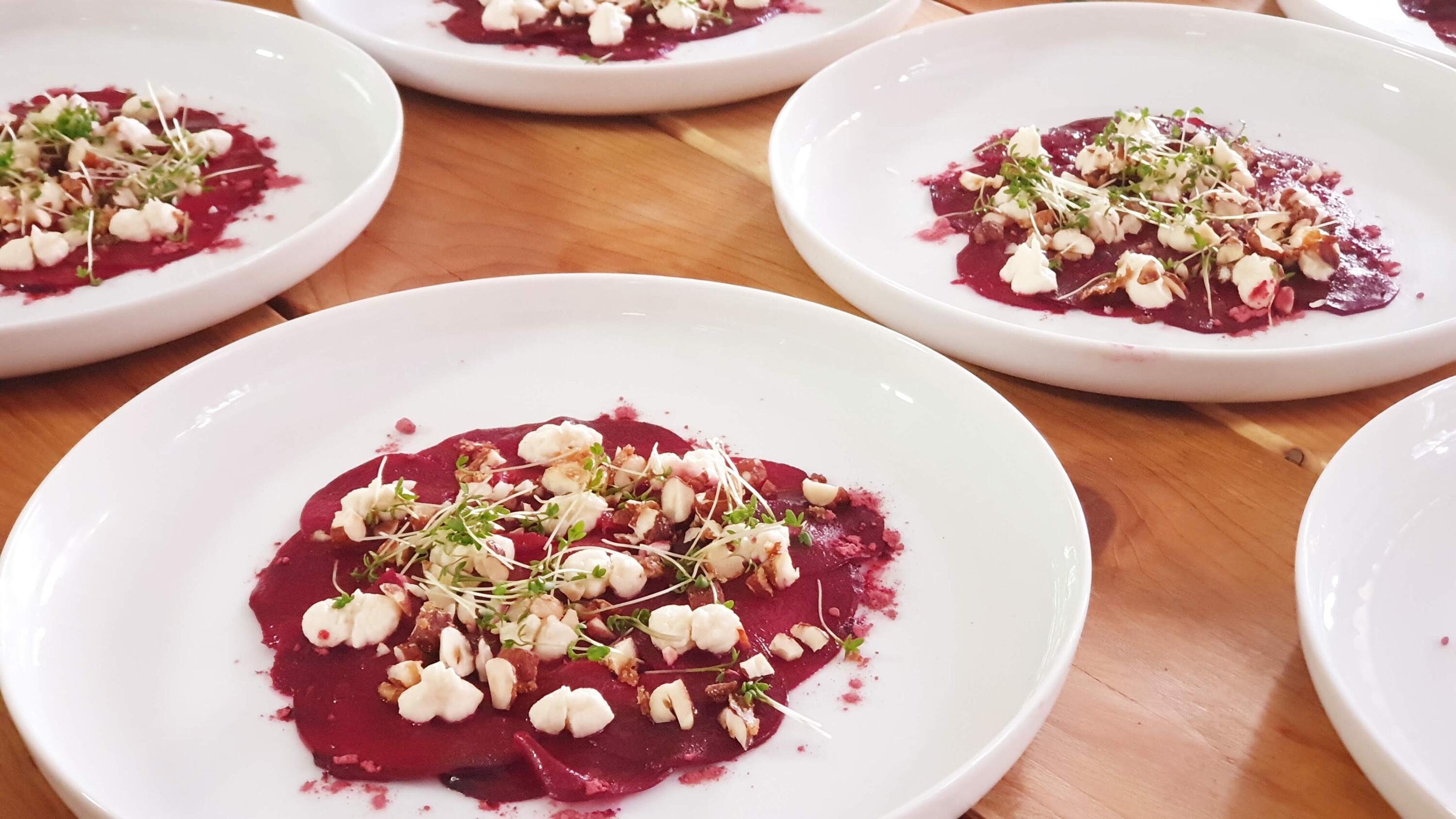 Weiße Teller mit rotem Rote Beete Carpaccio, garniert mit kleinen weißen Meerrettichstücken, karamellisierten Haselnussstücken und grünen Kresse-Sprossen, liegen auf einem Holztisch.