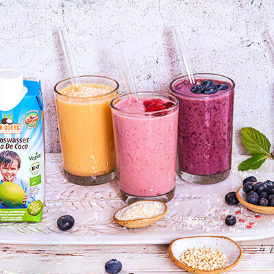 Detox Smoothies Drei bunte Smoothies in klaren Gläsern mit Strohhalmen stehen nebeneinander. Links daneben ist eine Packung Dr. Goerg Kokoswasser zu sehen. Vor den Gläsern liegen frische Beeren und ein Löffel mit Pulver.