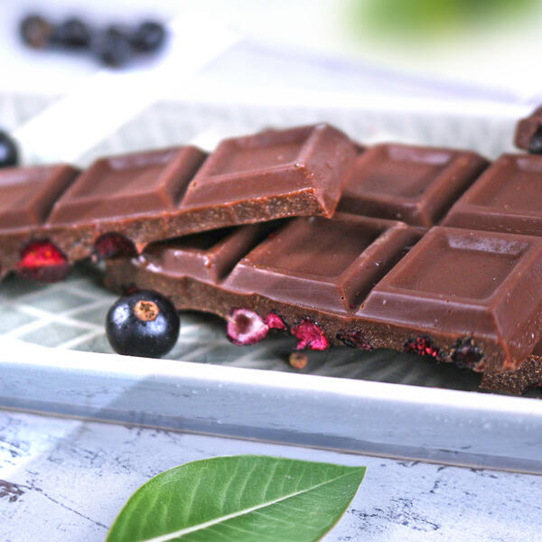 Feinherbe Cassis-Tahini Schokolade, roh-vegan Zwei Stücke feinherbe Schokolade mit getrockneten schwarzen Johannisbeeren liegen nebeneinander auf einem weißen Teller. Eine einzelne schwarze Johannisbeere liegt daneben. Im Vordergrund ist ein grünes Blatt zu sehen.