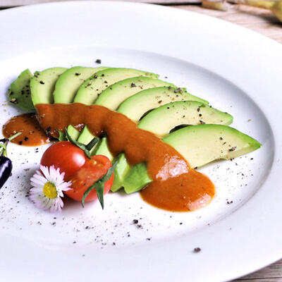 Süßsaure Chili-Soße, roh-vegan Auf einem weißen Teller sind mehrere Avocadoscheiben angerichtet, die mit süßsaurer roter Chili-Soße übergossen sind. Daneben liegen eine kleine Tomate und eine Blüte.