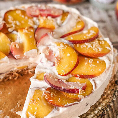 Pfirsich Raw Cake Roher Pfirsichkuchen mit cremigem weißem Belag wird serviert. Der Kuchen ist mit dünnen Pfirsichscheiben belegt und hat eine feuchte, nussige Basis. Eine Scheibe ist herausgeschnitten.