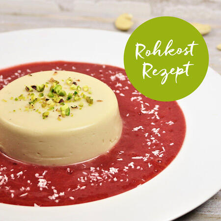 Weißes Panna Cotta mit gehackten Pistazien und Kokosraspeln steht auf roter Erdbeersoße auf weißem Teller. Grüner Kreis mit weißer Schrift Rohkost Rezept ist im Bild.