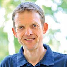 Peter E. Dreverhoff lächelt in die Kamera vor einem unscharfen, hellen Hintergrund mit grünem Blätterwerk.