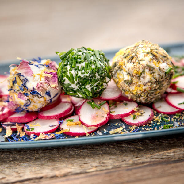 Mandelricotta von Boris Lauser Drei Mandelricotta-Kugeln liegen auf geschnittenen Radieschen. Die Kugeln sind mit Essblüten, Schnittlauch und Gewürzen garniert. Das Gericht wird auf einem dunklen Teller serviert.