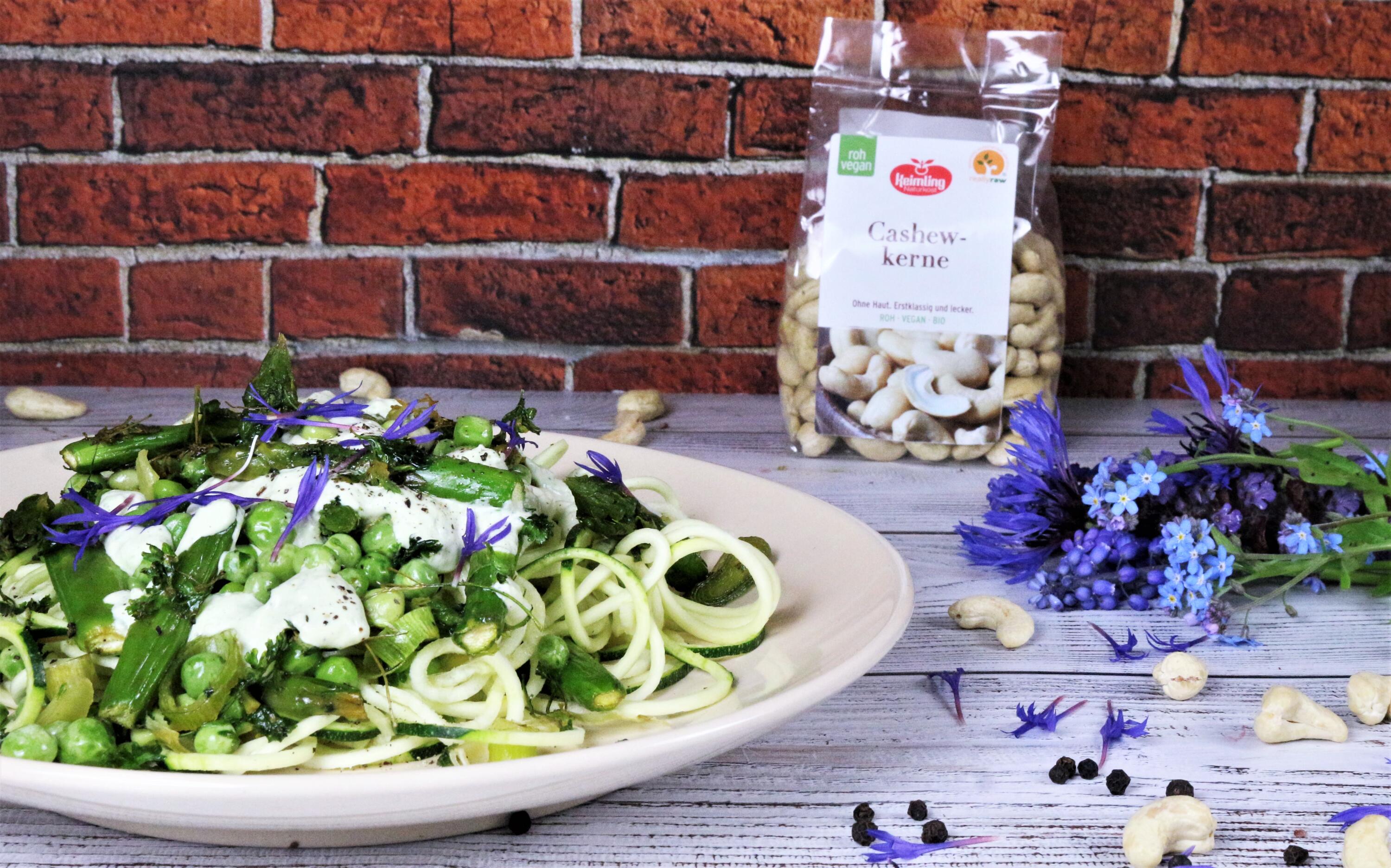 Ein Teller mit Zucchinispaghetti, Zitronenspargel, Erbsen und weißer Kräutercreme, garniert mit violetten Blüten. Daneben steht eine Packung Cashewkerne und ein Strauß blauer Blumen auf einem Holztisch.