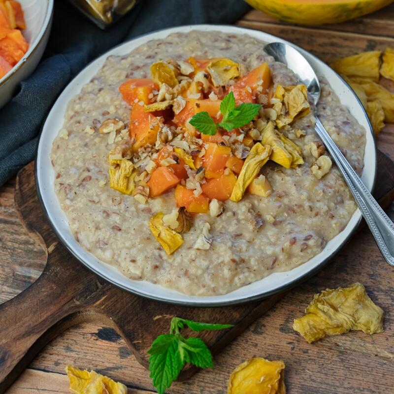 Cremiges Bananen-Porridge mit Mangostücken und Papaya Ein Teller cremiges Bananen-Porridge wird mit frischen Mangostücken, Papaya, gehackten Nüssen und einem Minzblatt garniert. Ein Löffel liegt am Tellerrand.