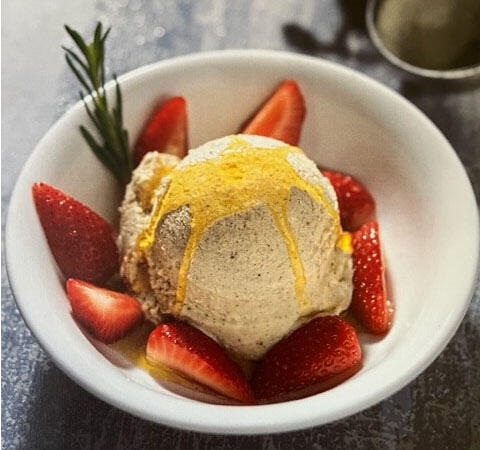 Cashew-Eis_Rezept_Keimling Eine Kugel veganes Vanille-Eis mit goldener Sauce beträufelt. Die Eis-Kugel liegt in einer weißen Schale, umgeben von frischen Erdbeerhälften und einem Rosmarinzweig.