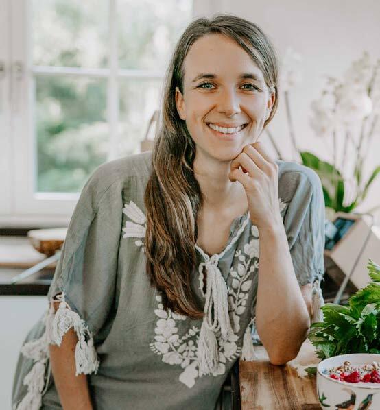 Ute Ludwig sitzt lächelnd in einer Küche und stützt ihren Kopf mit der Hand. Sie trägt ein olivgrünes, besticktes Oberteil und posiert vor einem Fenster mit Pflanzen im Hintergrund.