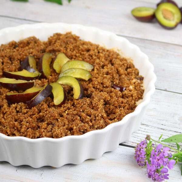 Zwetschgencrumble mit Vanille-Zimtcreme, roh-vegan Ein Weißes, rundes Keramik-Backform mit einem braunen Zwetschgencrumble, der mit mehreren geschnittenen Zwetschgenstücken in der Mitte garniert ist. Daneben liegt eine lila Blüte und im Hintergrund halbierte Avocados auf einem weißen Holztisch.