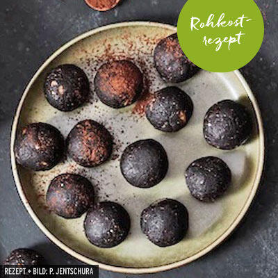 Runde, dunkle Energiebällchen liegen auf einem hellen Teller. Einige sind leicht mit Kakaopulver bestäubt. Ein grüner Aufkleber zeigt den Text Rohkost rezept.