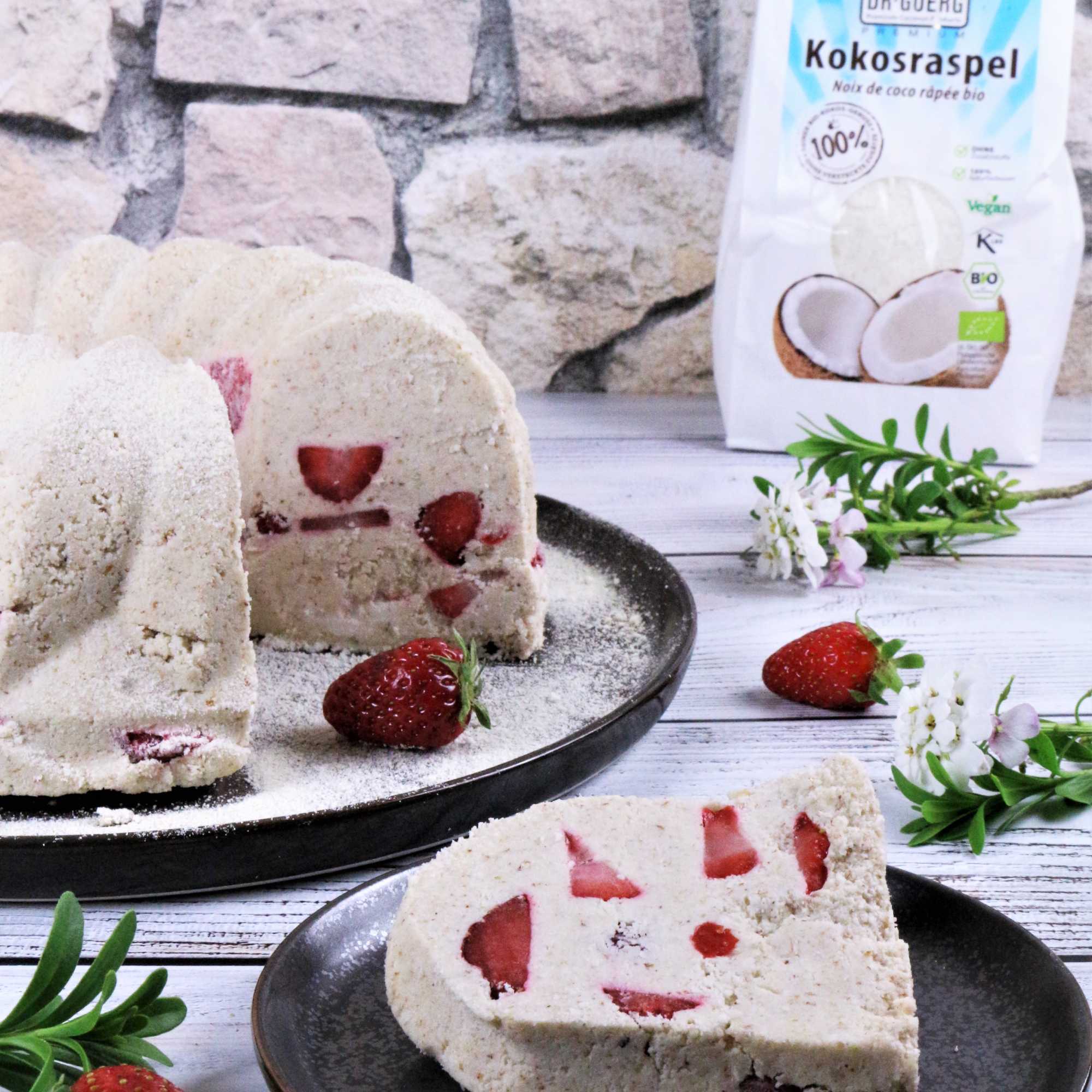 Leichter Gugelhupf mit Erdbeeren, roh-vegan Leichter Rohkost-Gugelhupf mit frischen Erdbeeren auf einem schwarzen Teller. Ein Stück Gugelhupf mit sichtbaren Erdbeerstücken liegt auf einem kleinen grauen Teller. Im Hintergrund ist eine Packung Kokosraspel zu sehen.