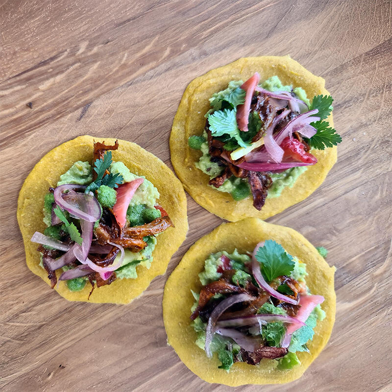Rohkost-Rezept: Tacos al Hongo Pibil von Boris Lauser Drei vegane Tacos auf Holzuntergrund mit gelbem Mais-Tortilla, grüner Guacamole, gebratenen Pilzen, roten Zwiebeln, Koriander und Pickles belegt.