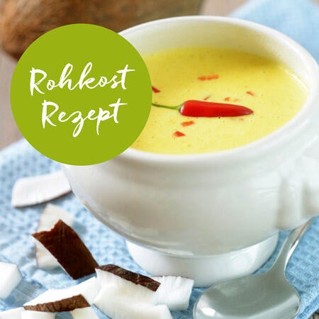 Weiße Schale mit gelber Rohkostsuppe, garniert mit rotem Chili, steht auf blau-weiß kariertem Tuch. Daneben liegen Stücke frischer Kokosnuss und ein Löffel.
