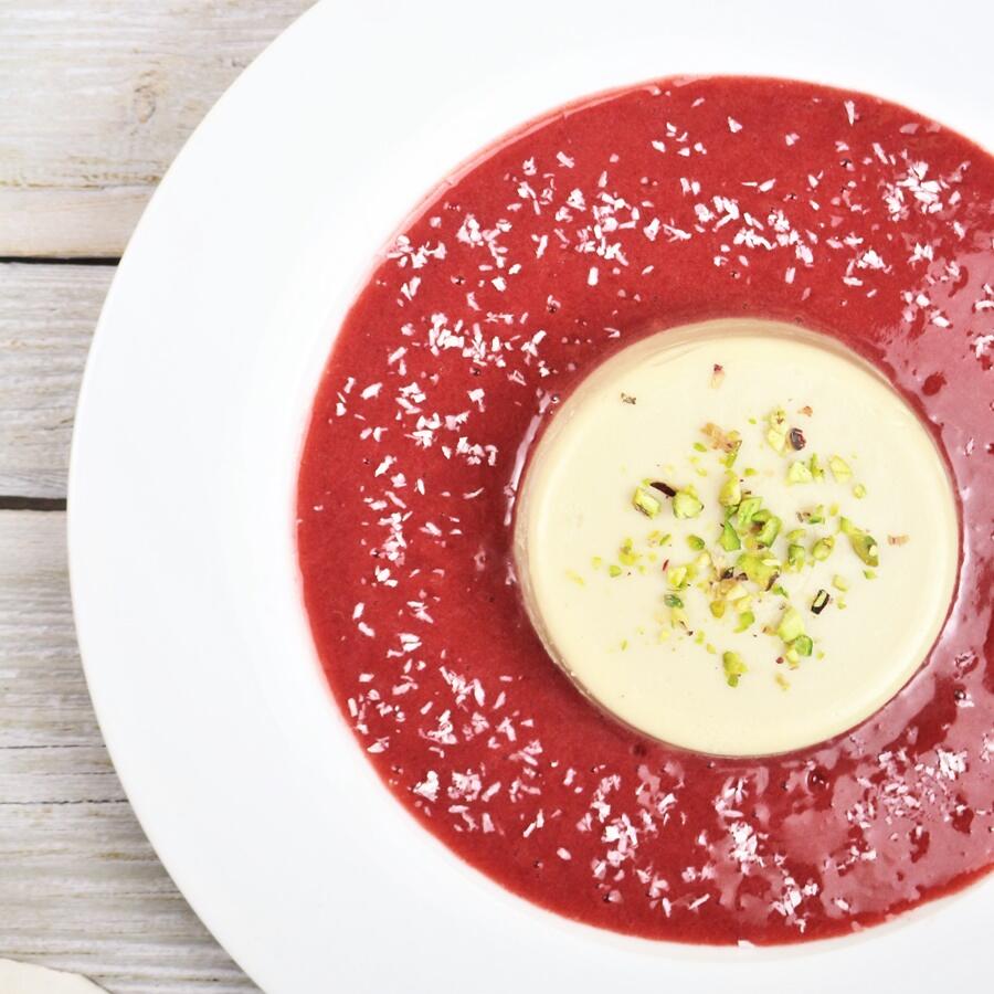 Ananas Panna Cotta mit Erdbeersoße, roh-vegan Weiße Ananas-Panna-Cotta mit gehackten Pistazien obenauf steht in der Mitte eines weißen Tellers. Umgeben wird sie von roter Erdbeersoße, die mit Kokosraspeln bestreut ist.