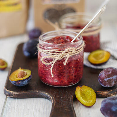 Zwetschgen Chutney Ein Glas Zwetschgen-Chutney mit Juteband dekoriert steht auf einem Holzbrett. Daneben liegen halbierte und ganze Zwetschgen. Ein Löffel steckt im Chutney.