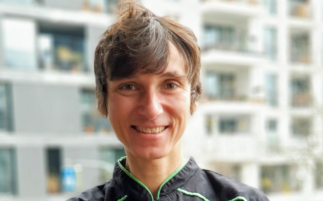 B. Lauser, Rohkost- und Pflanzenkoch aus Berlin, lächelt in die Kamera. Er trägt eine schwarze Kochjacke mit grüner Naht. Im Hintergrund verschwommene Gebäude.